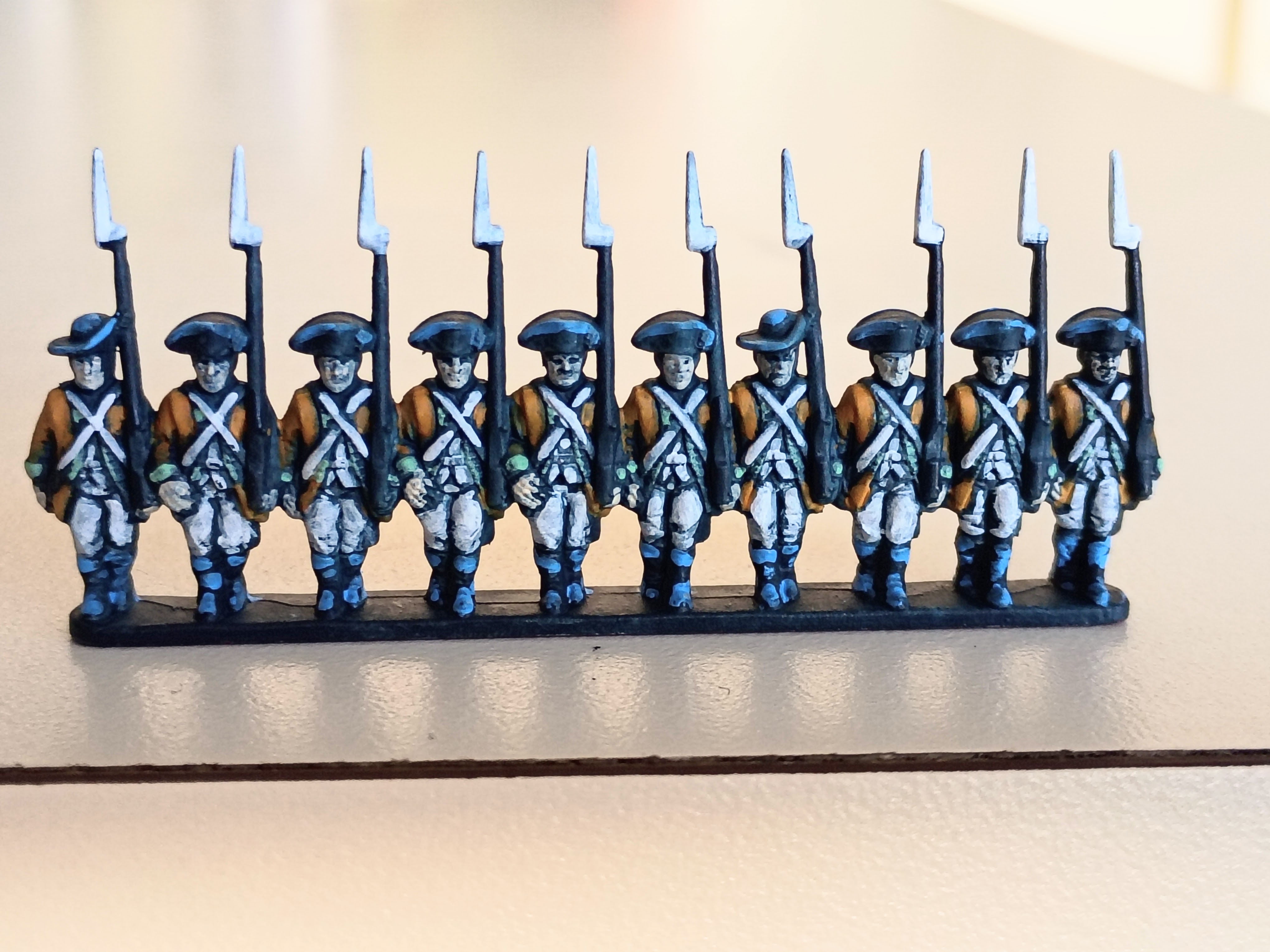 A line of Warlord Games Epic Battles Revolution line infantry painted hastily. They wear yellow-orange coats, white shirts and vests, white breeches, and feature a light green trim. The base color, which shows through as the shadow throughout the miniature but also appears as a substitute for brown, is a deep sea blue or dark teal. The tricorns, gaiters, and footwear, which would normally be black, is only highlighted in light blue instead.